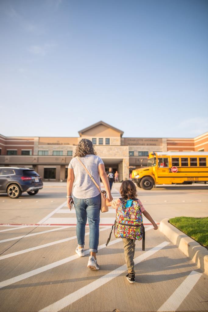 A white mom and daughter hold hands. We see their backs as they walk toward a school with a school bus in front of it.