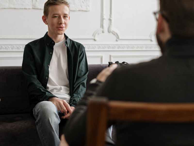 A white teen looks somber as he speaks to his male therapist. We see the therapist only from behind. Both are sitting.
