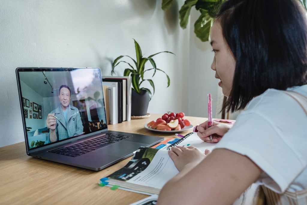 An Asian girl sits at her desk with an open book in front of her. Behind the book is an open laptop with her Asian father. They are talking on a video call.