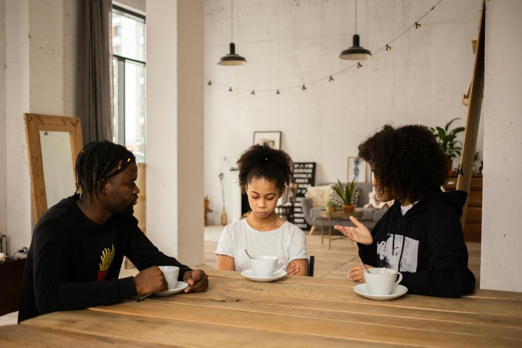 A mother, father, and daughter of color sit at a kitchen table. The parents are explaining something to the child. The girl is looking down as if she is disappointed.