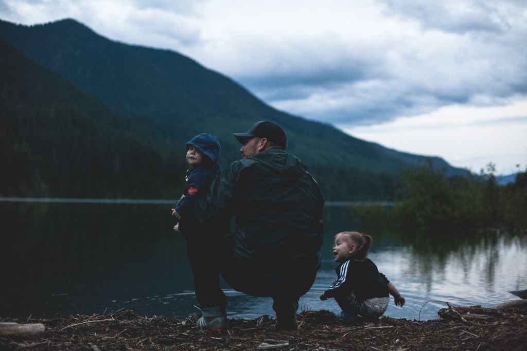 A white father and his two children are facing a lake with a mountain the distance. We see them from behind. The father is kneeling , as is the toddler next to him. The dad has a baby on resting on his knee.