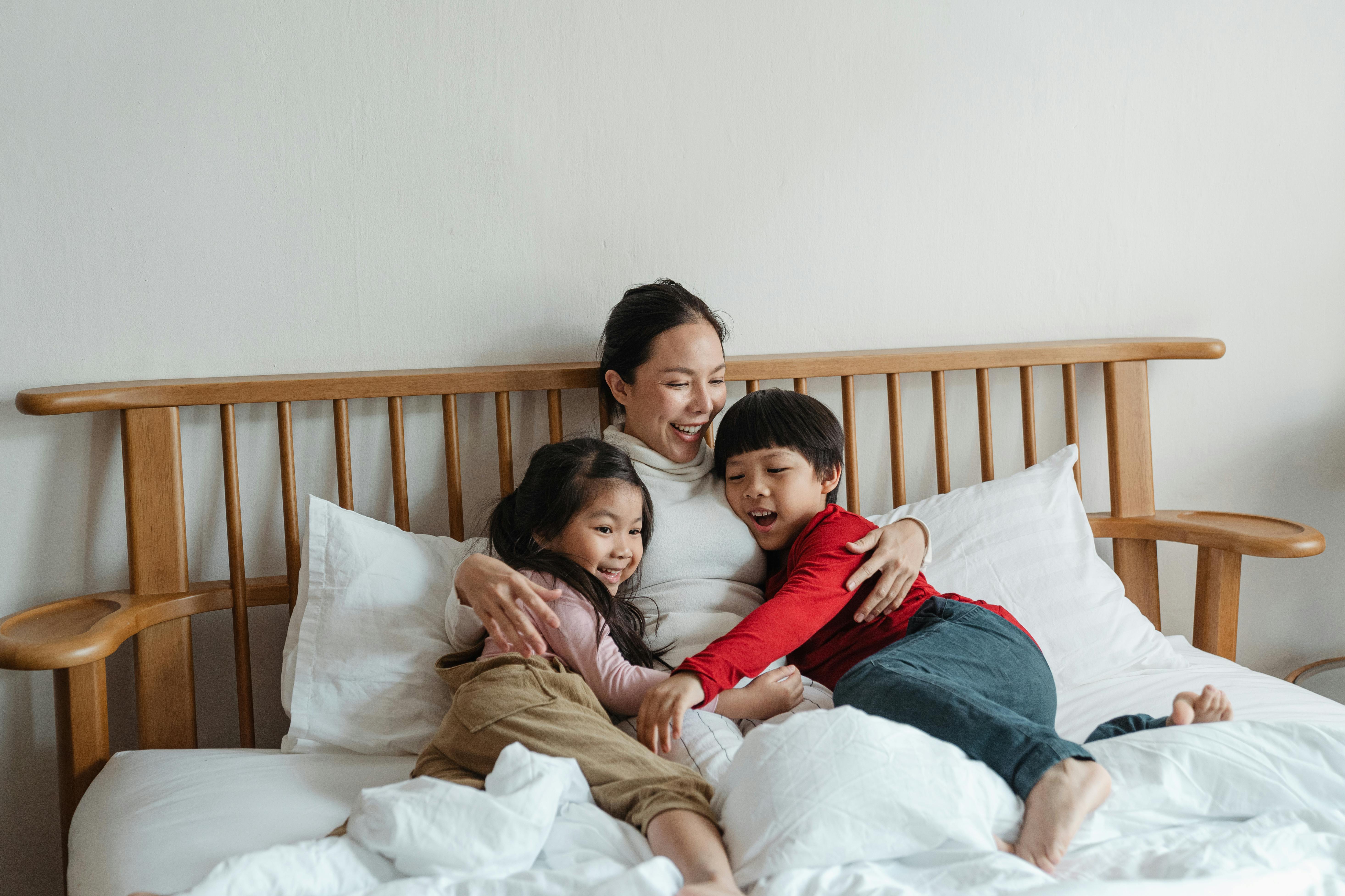 As Asian mother smiles as she hugs a son on one side of her and a daughter on the other. The mother and two young children are sitting on a bed with white bedding.