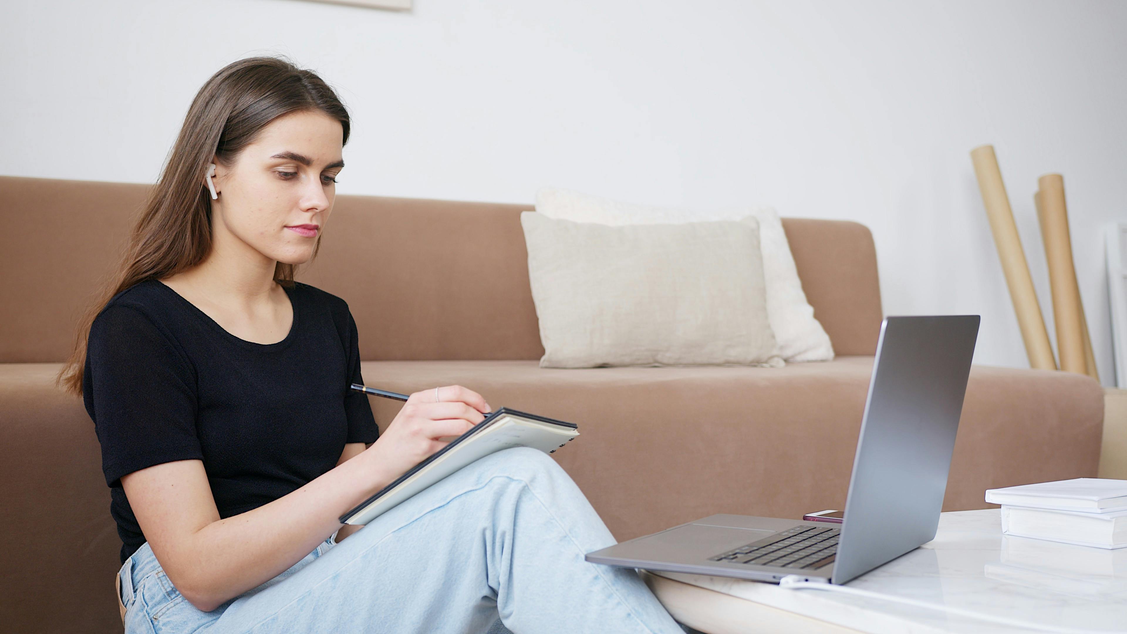A white girl sits on the floor with a notebook resting on her bent knee. She is leaning against a tan couch. She is writing in her notebook with a computer open in front of her.