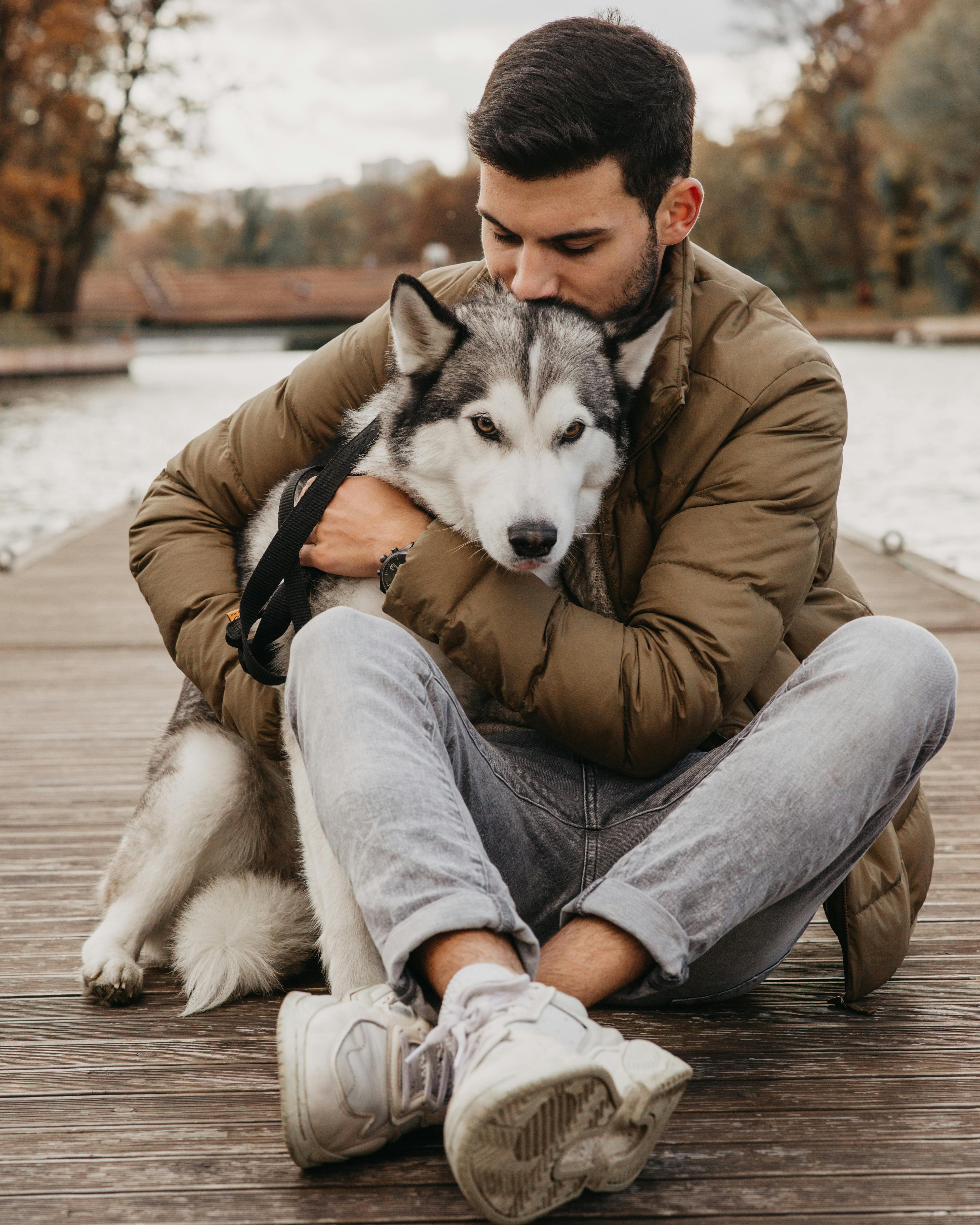 A man sits on the ground as he hugs a grey and white husky dog.