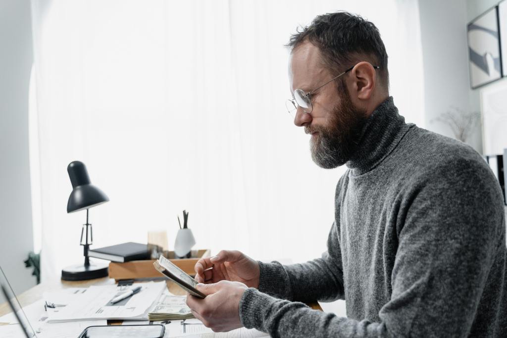 A white man in a turtleneck is looking at papers as he sits at a desk.
