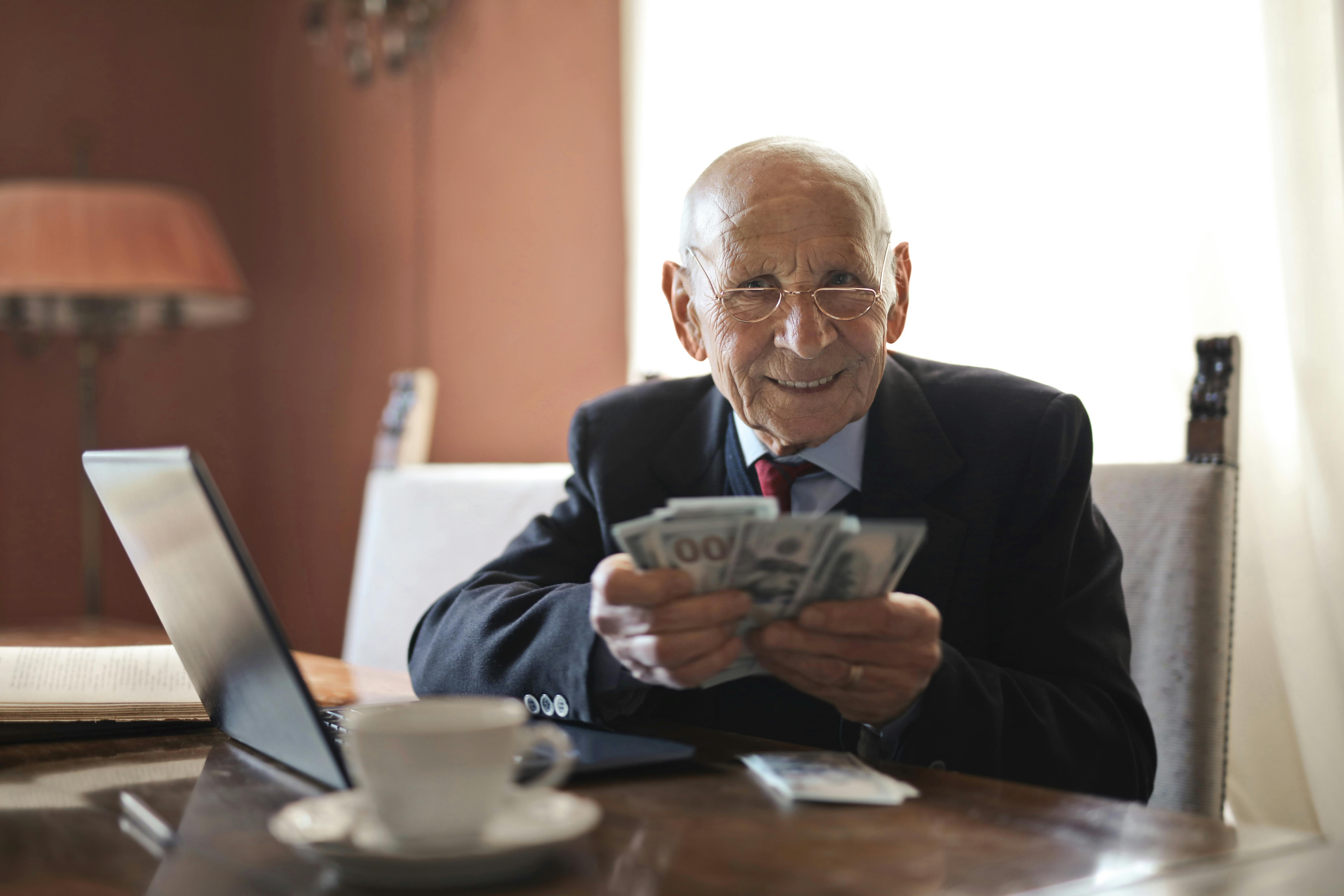 An elderly white man is dressed in a suit and sitting at a desk. He is fanning out hundreds of dollars and smiling.