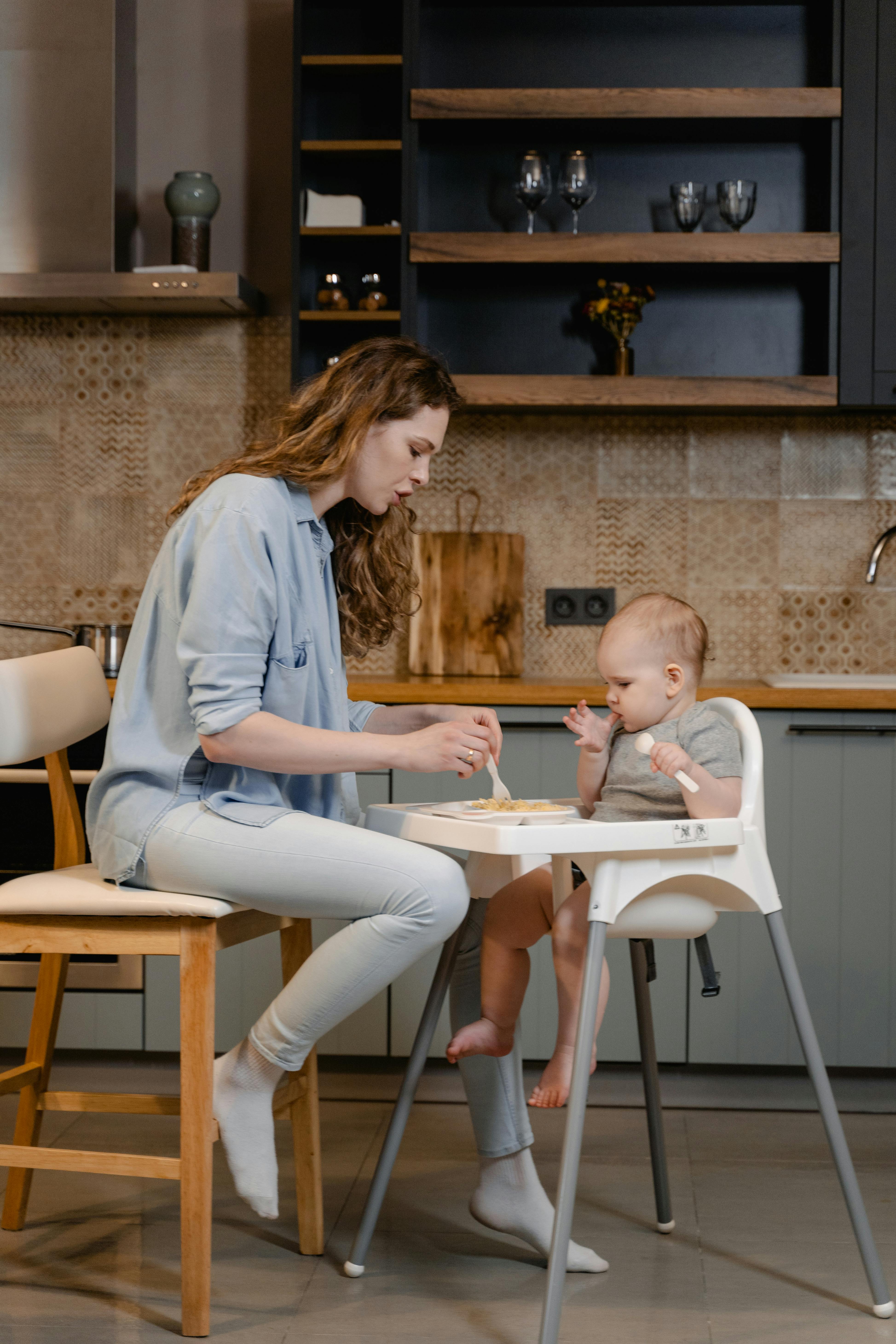 A white woman sits in a chair. She is feeding her baby who is sitting in a high chair.