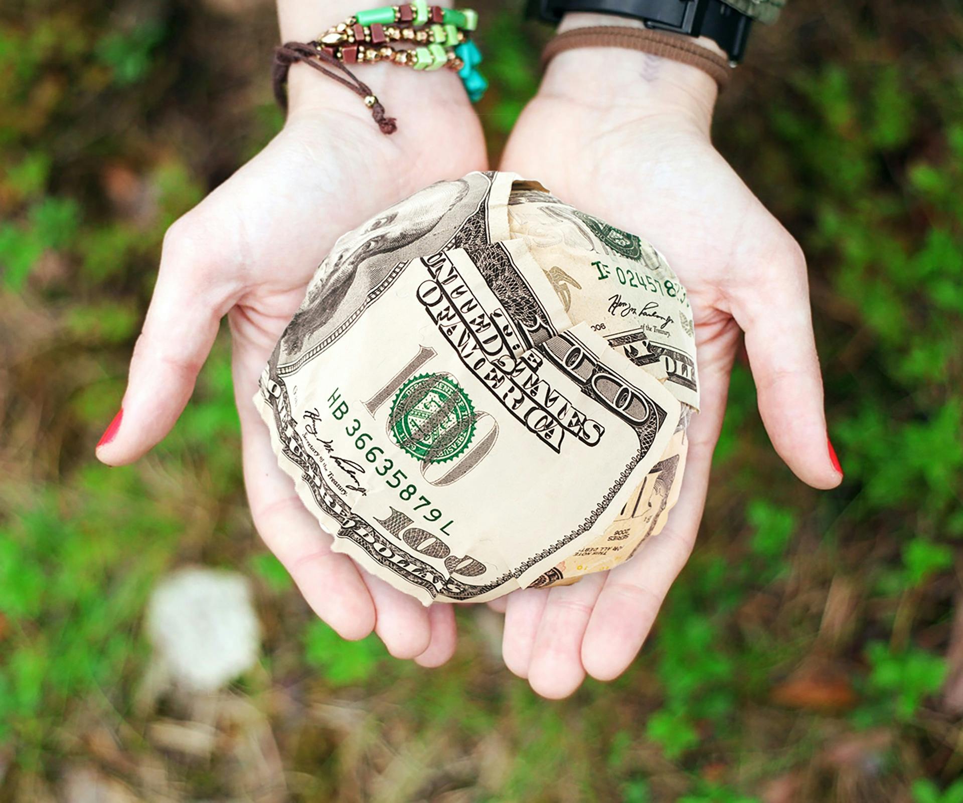 Two white hands hold a ball of cash. Beneath the hands is a forest floor.