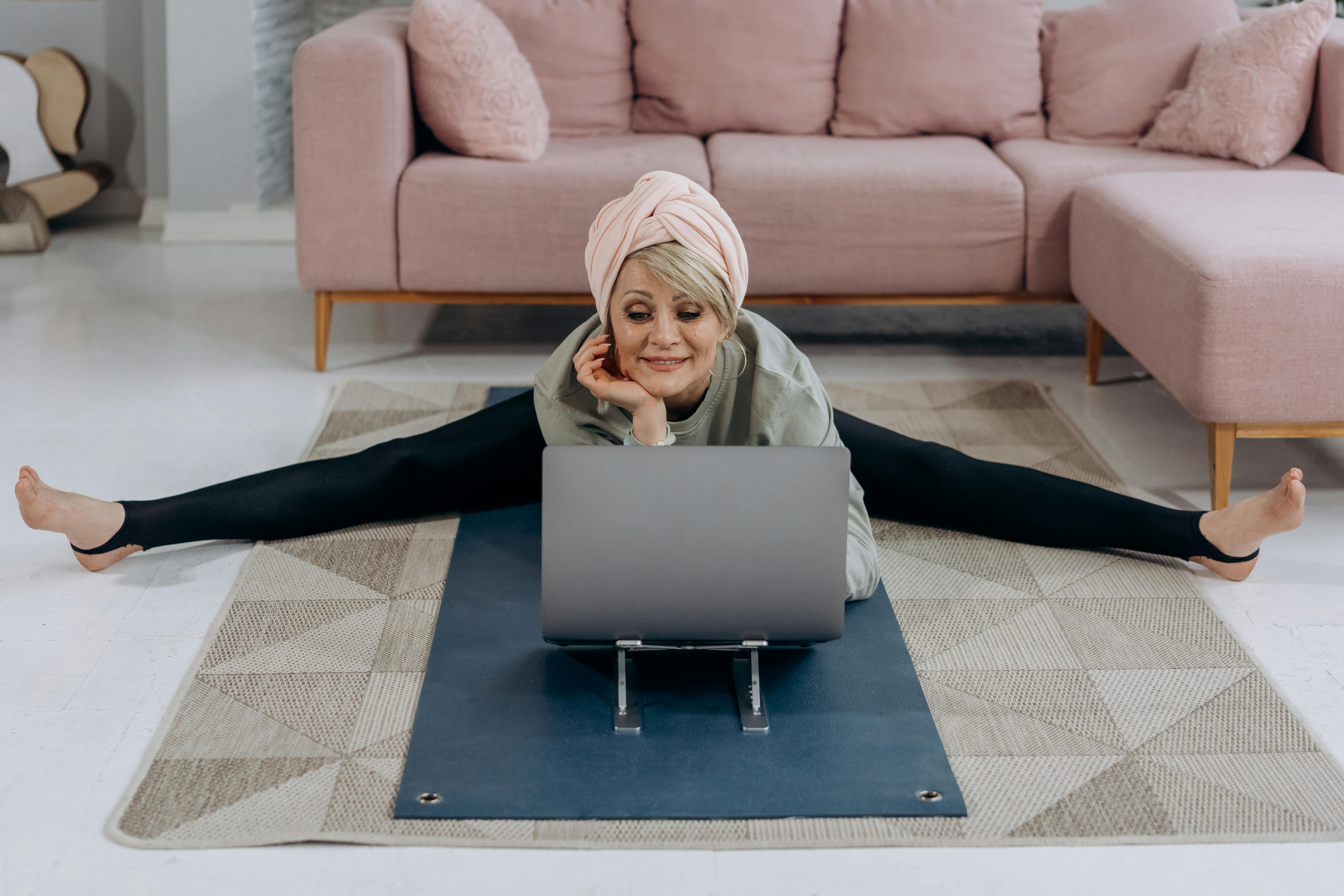 A middle-aged white woman is stretching on a living room floor as she looks at her laptop.