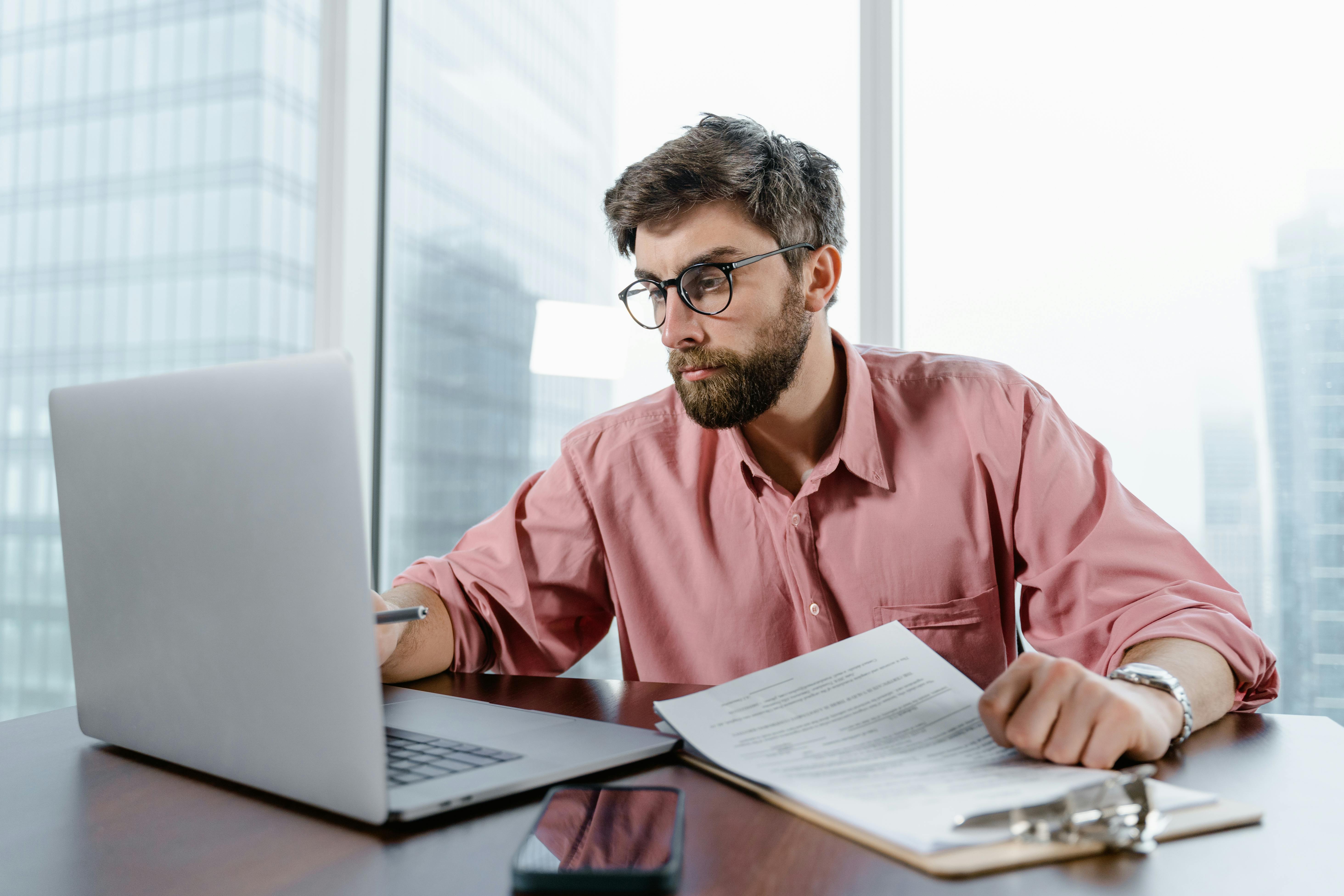 A white man sits at a desk and is looking at a computer and some papers on a clipboard.