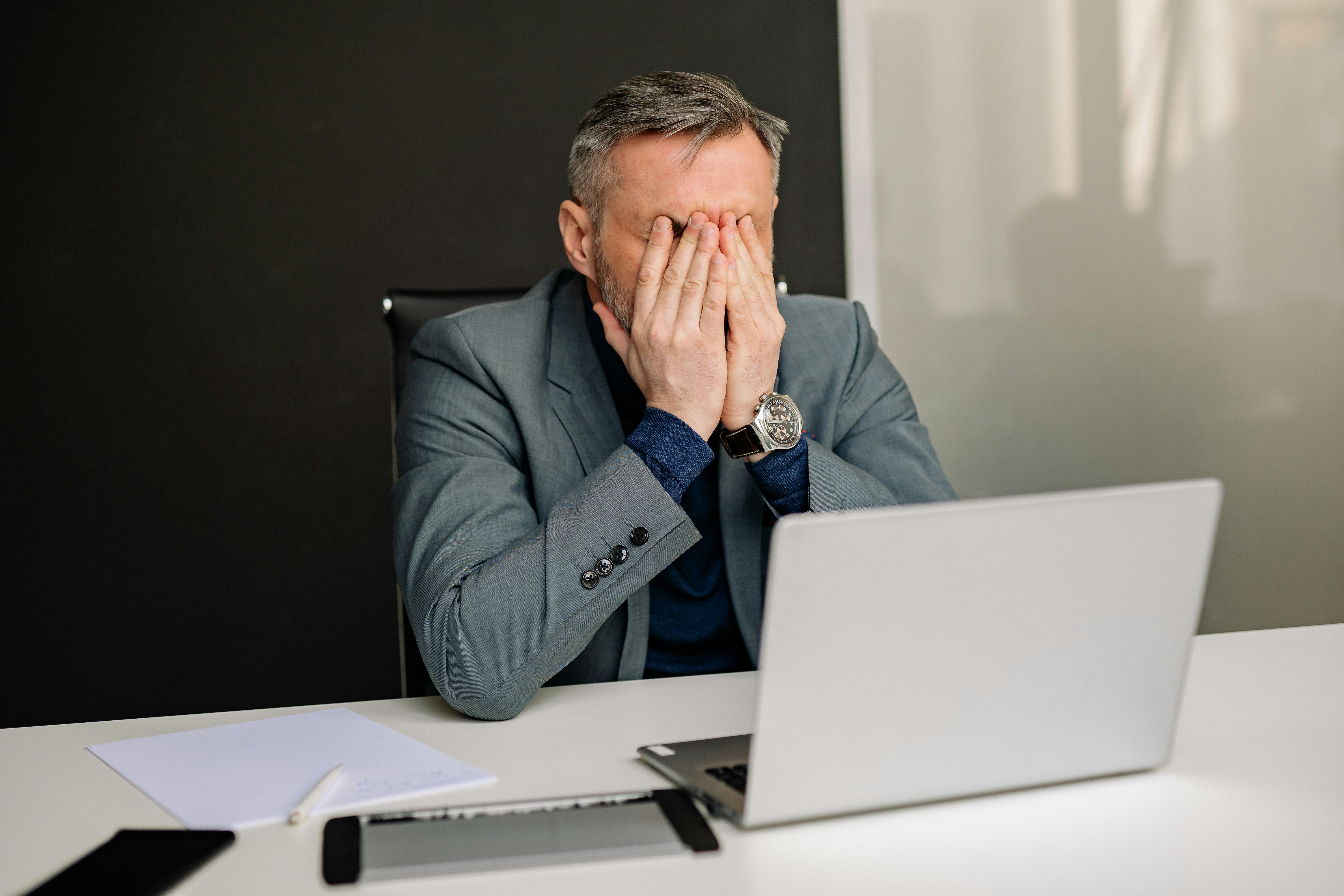 A white man is sitting at a desk with a computer in front of him. His hands are pressed into his eyes in frustration.