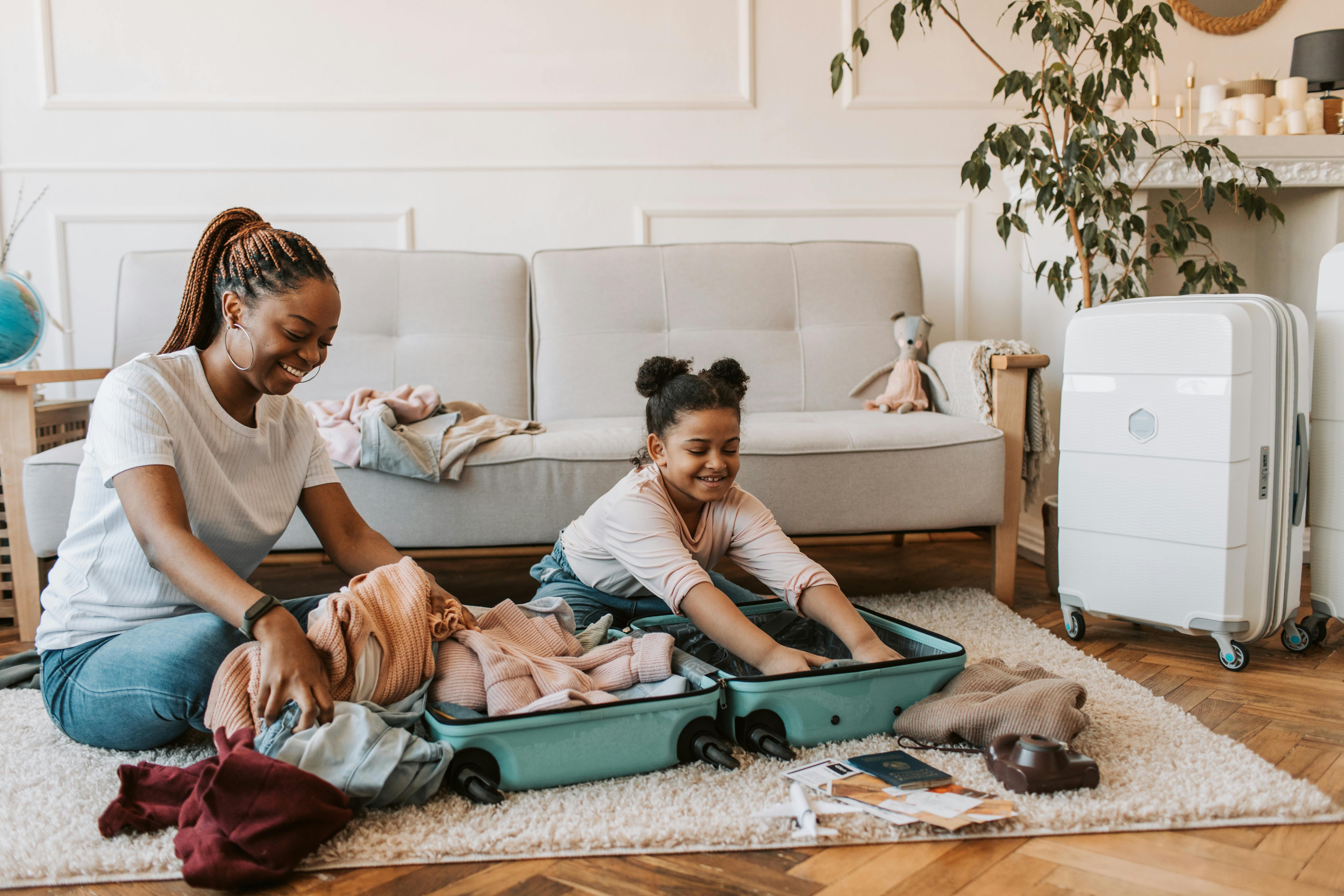 A black mom is sitting with her daughter on the living room floor. There is an open suitcase in front of them. The mom is helping the daughter pack to see her dad for visitation.