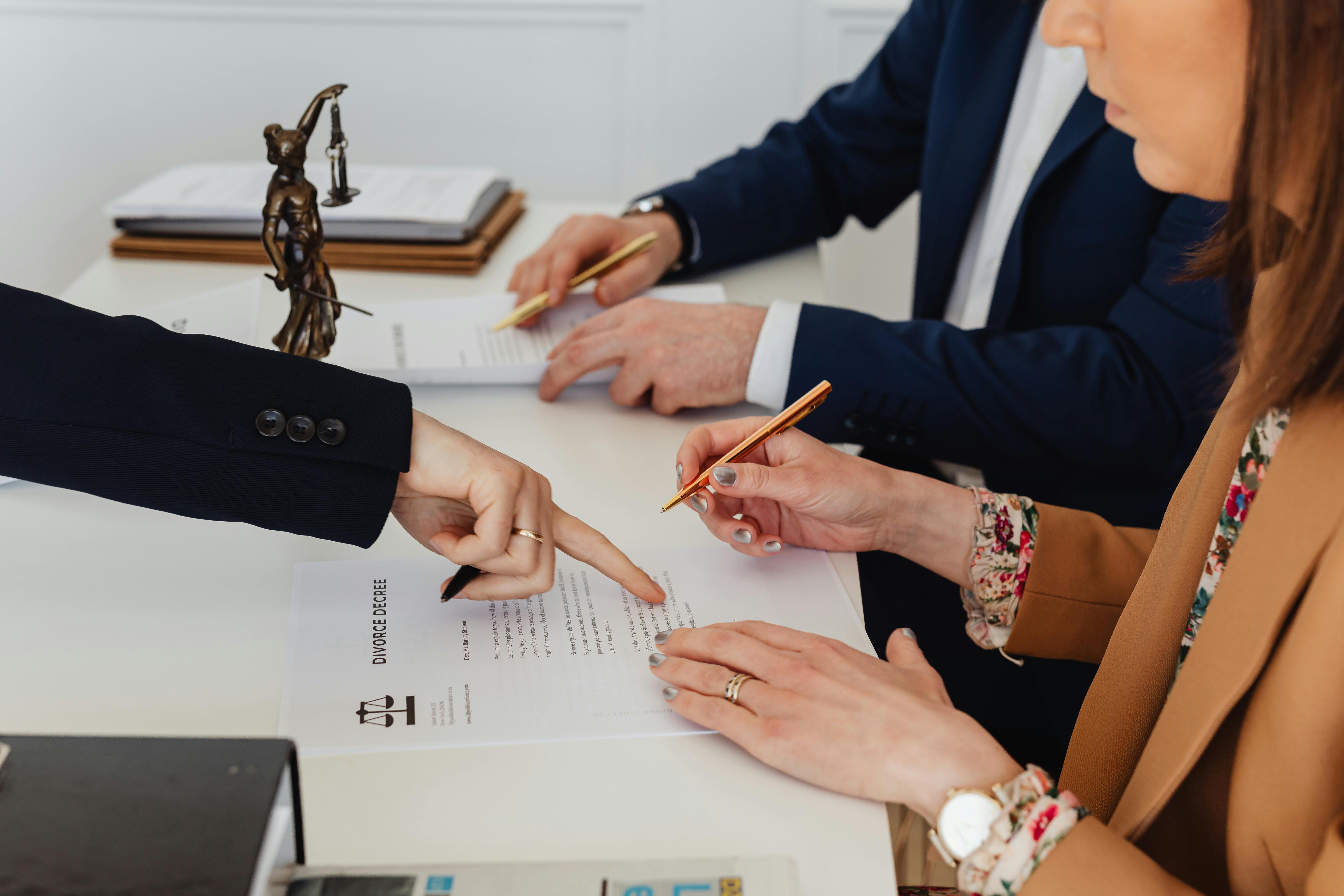 A close up photo of three sets of hands. One set is behind a desk and pointing to papers on the desk. The other two sets belong to a married couple waiting to sign divorce papers.