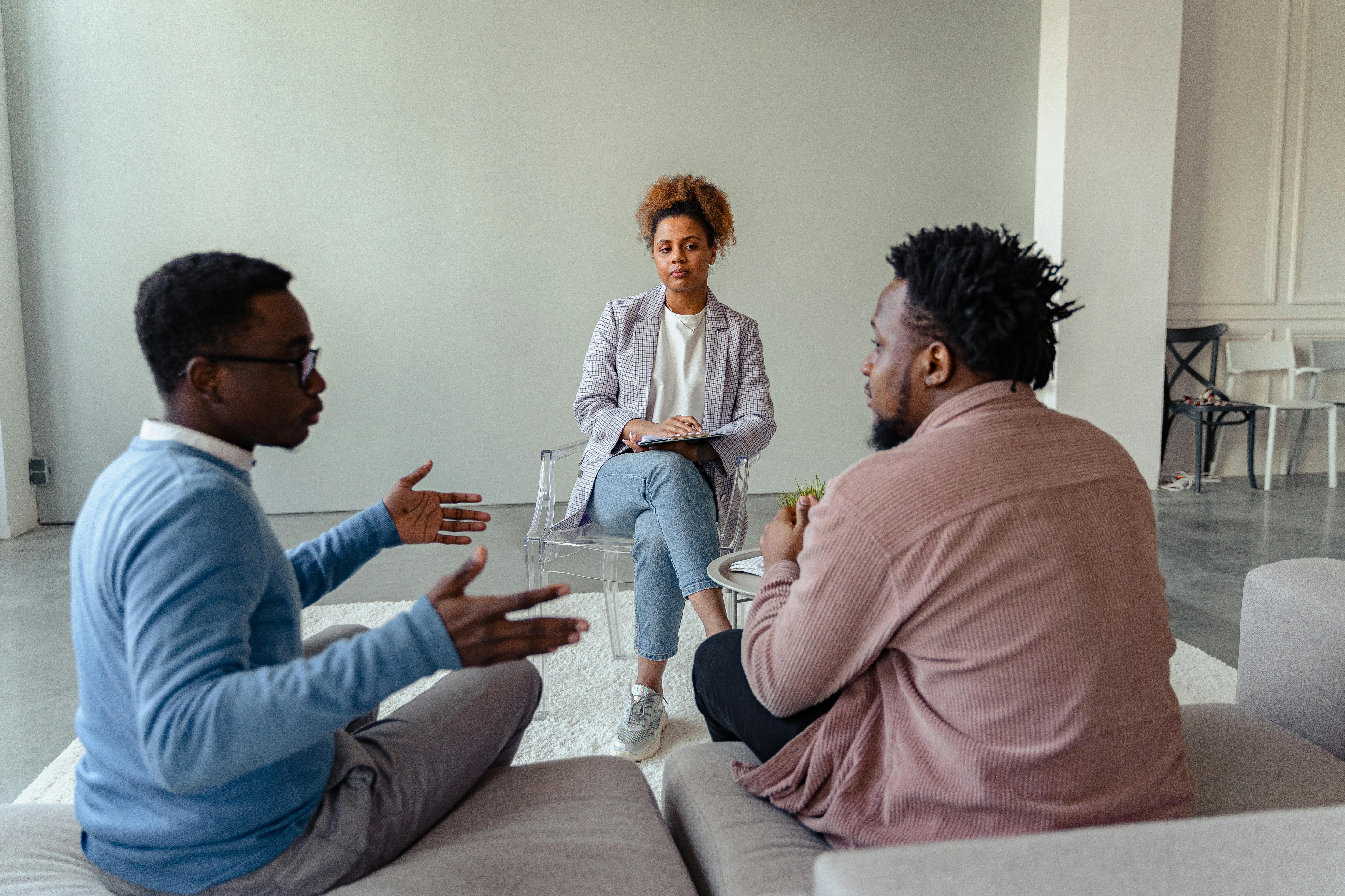 A black LGBTQ couple is sitting on a couch with a black mediator across from them in a chair. They are in mediation to settle their divorce.