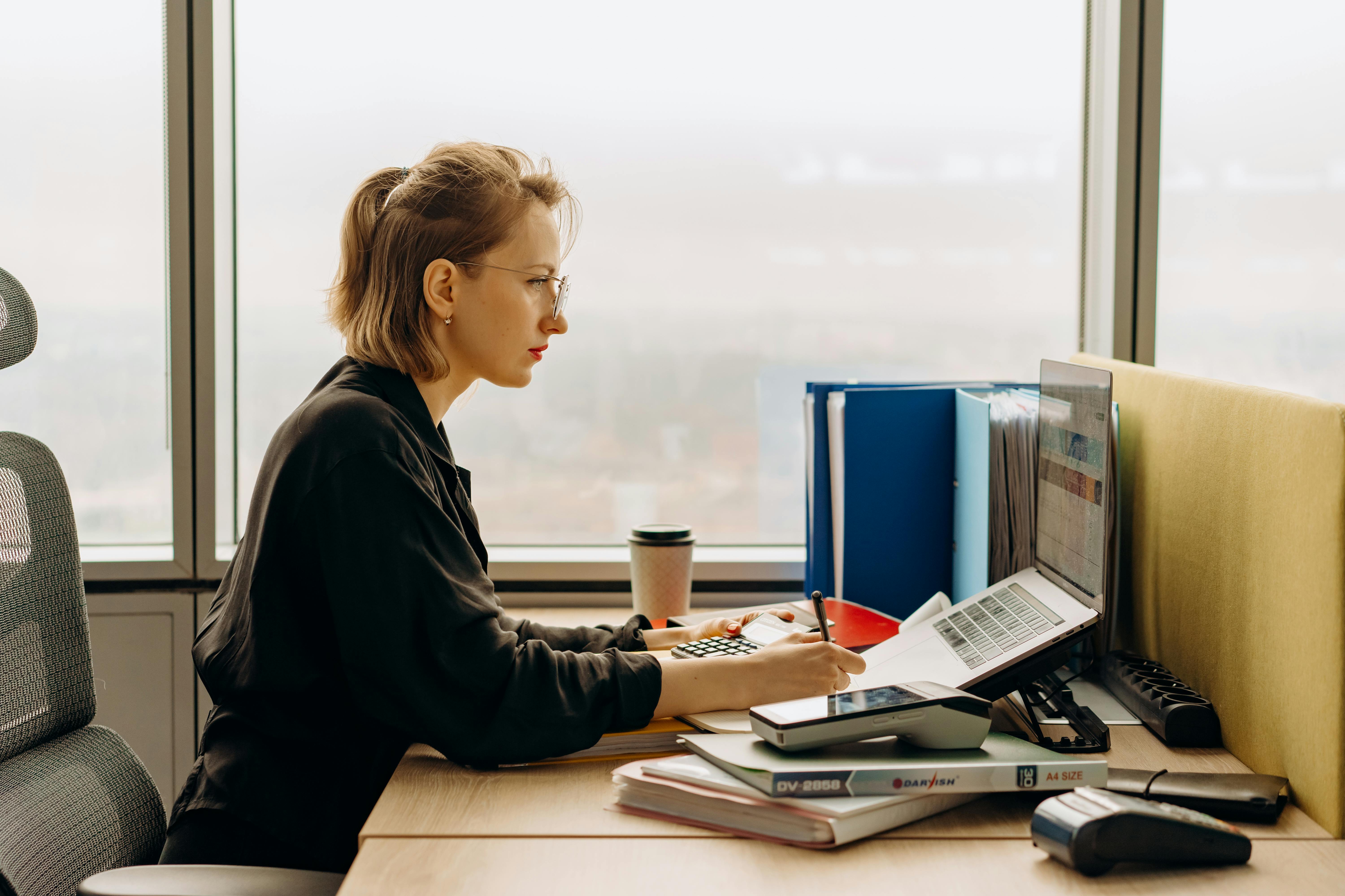 A white woman sits at a desk with a city skyline out the window to her side. She is working at a computer.