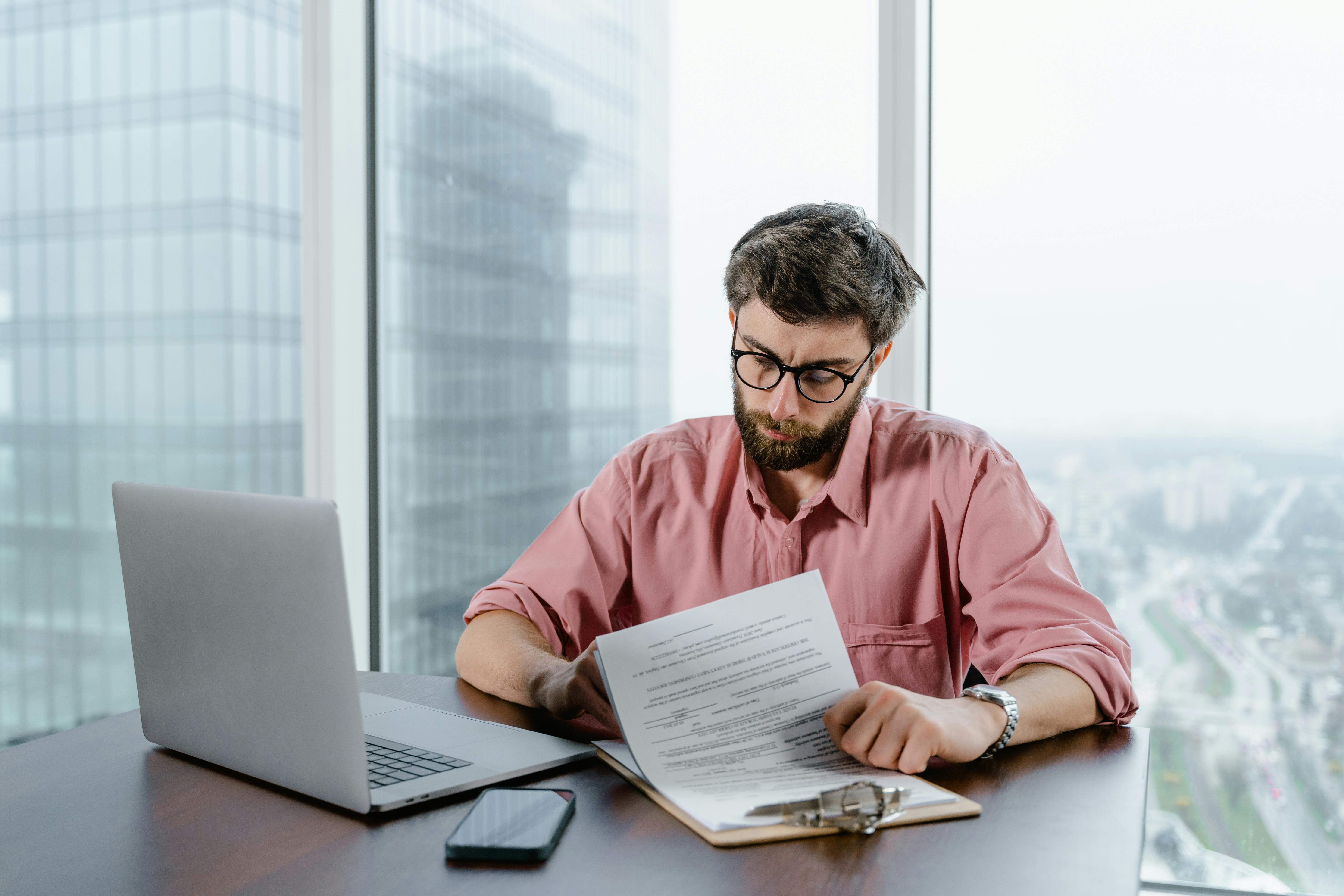 A young white man is sitting in his high rise office in front of a large window. He is signing divorce papers at his desk.