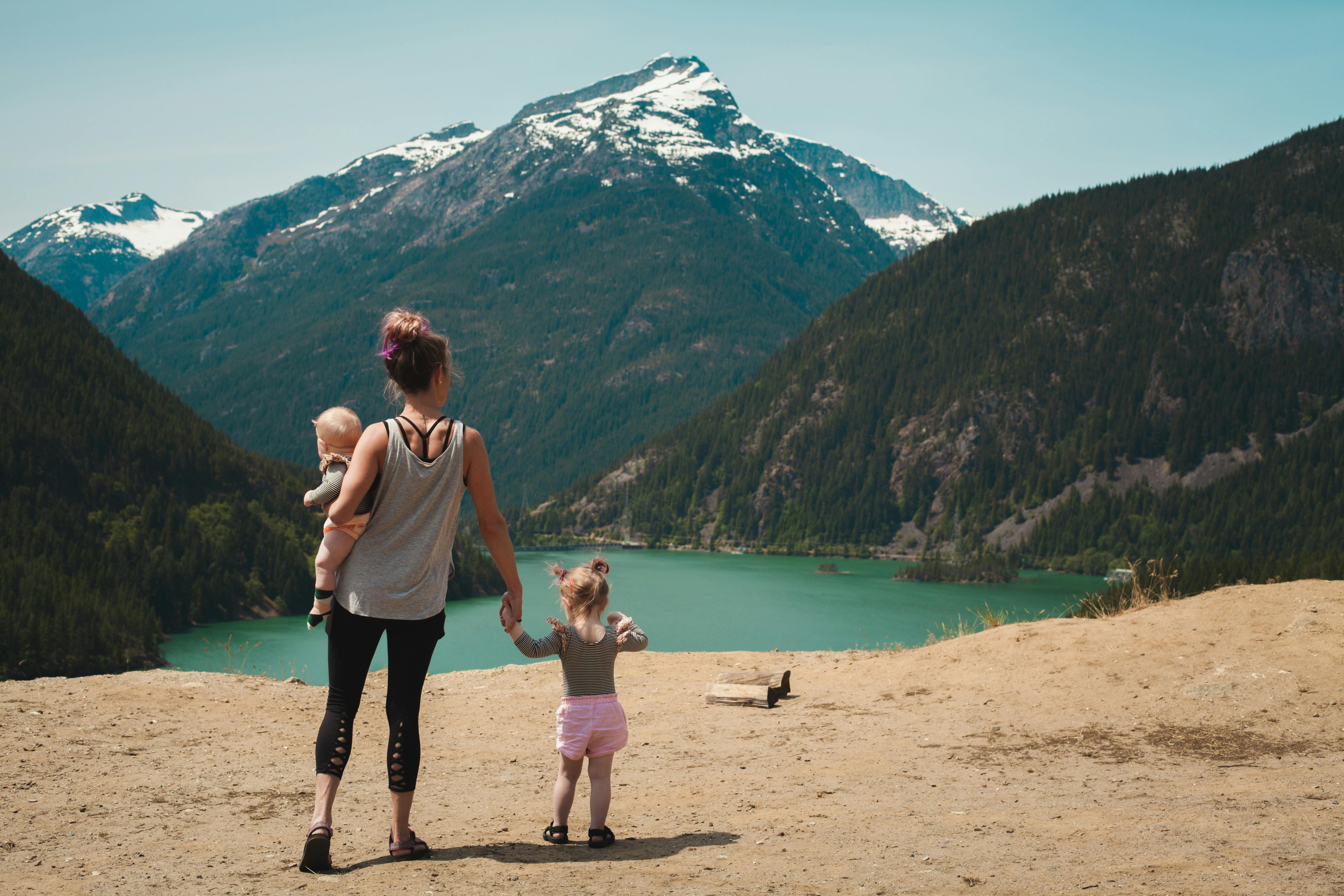 A mom is holding her baby and holding her young daughter's hand. They're all facing a mountain and lake. We see them from behind.