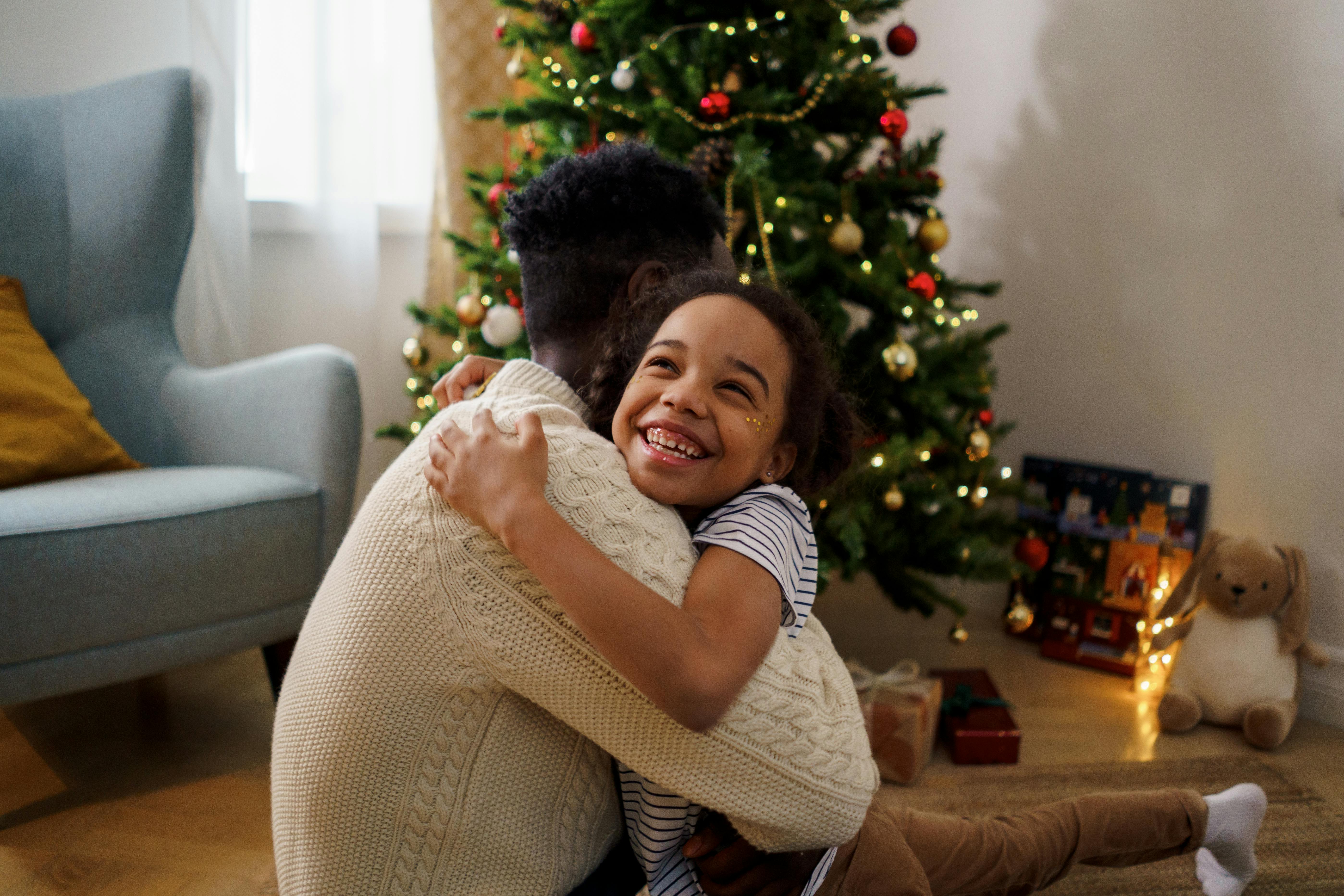 A black dad is hugging his daughter in front of a Christmas tree.