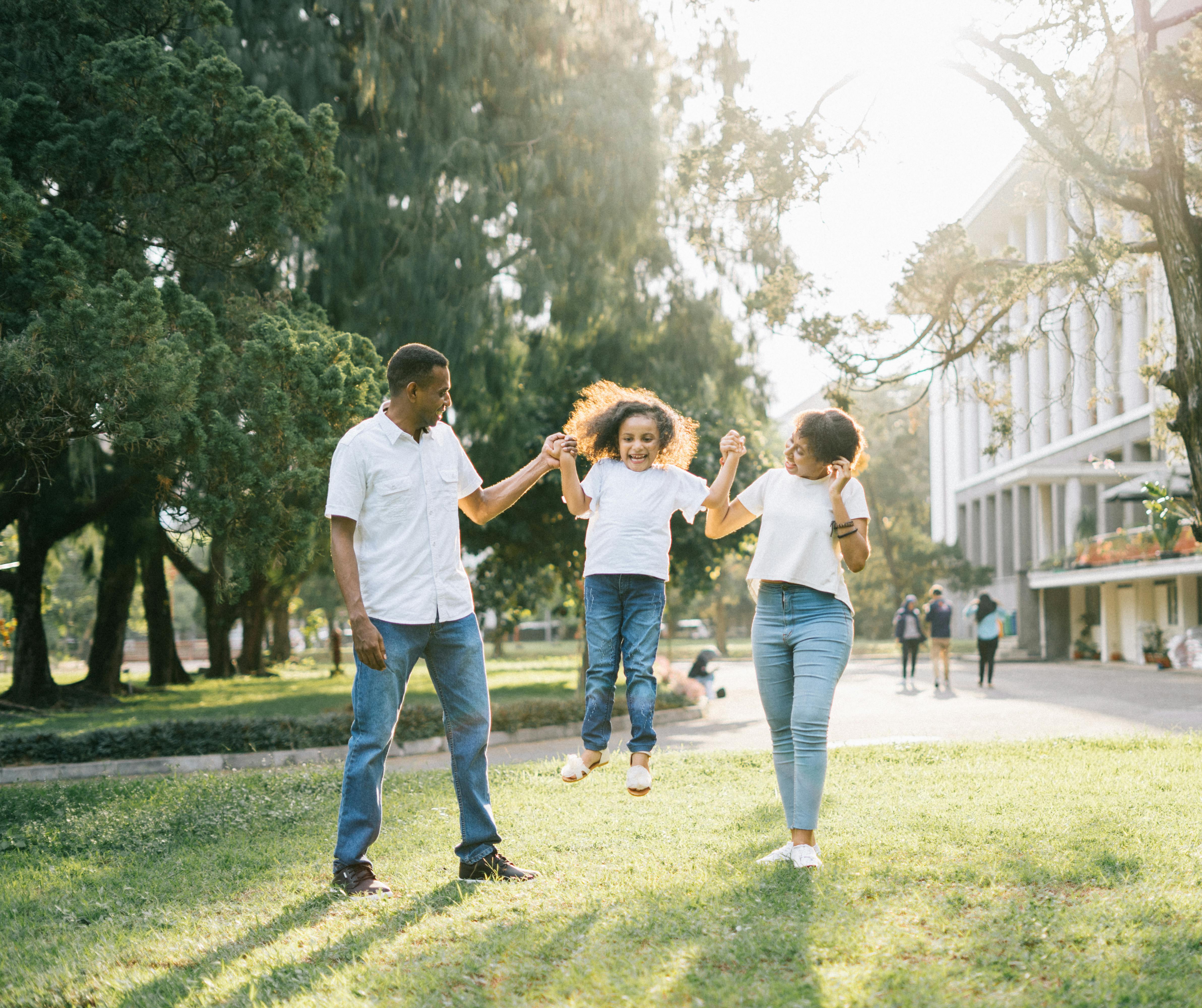 A black dad, black mom, and black daughter are all in white shirts and jeans. The parents are lifting their daughter off the ground and swinging her.