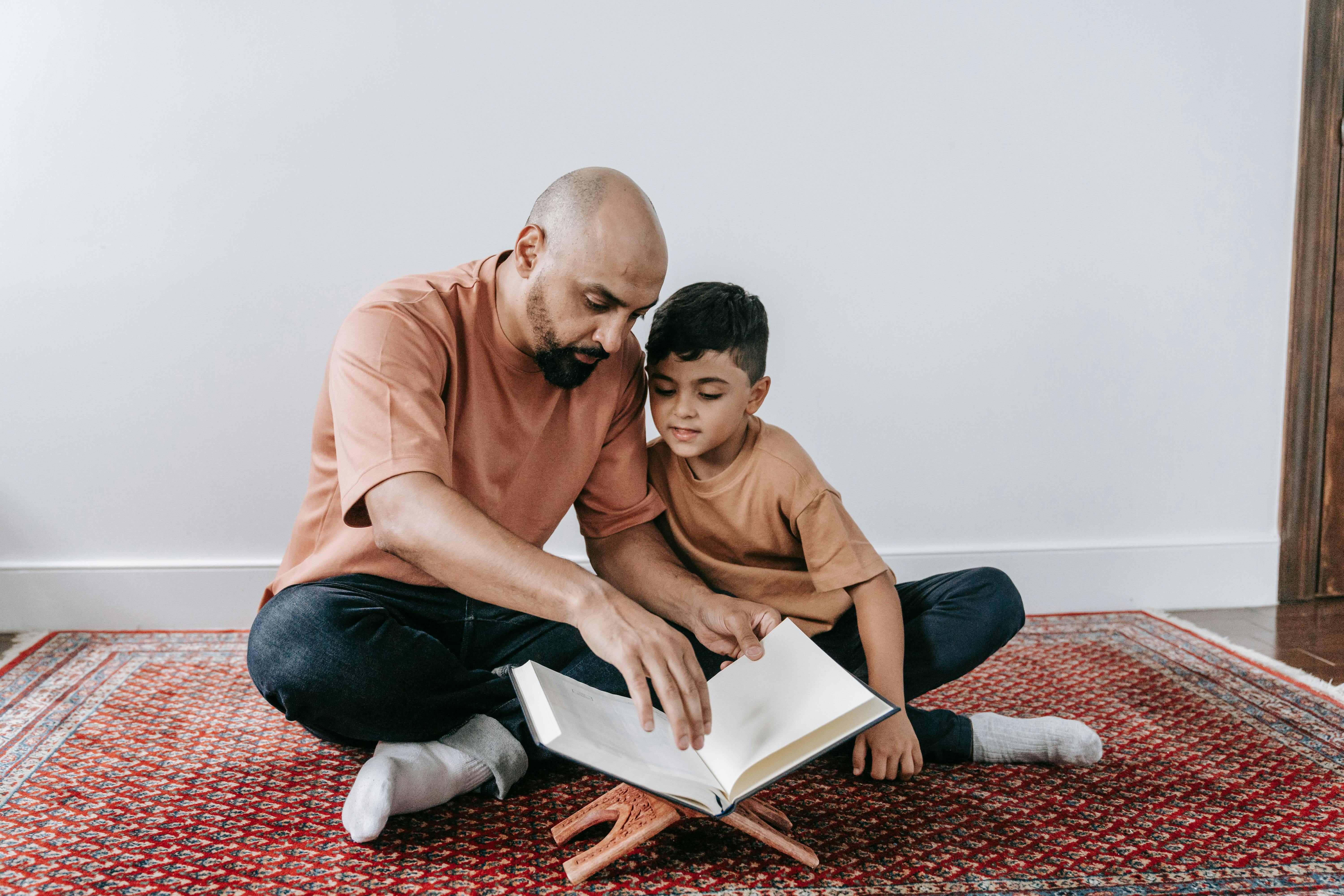 A father of Middle Eastern heritage sits on the floor with his son. They're reading a book together on a book stand.