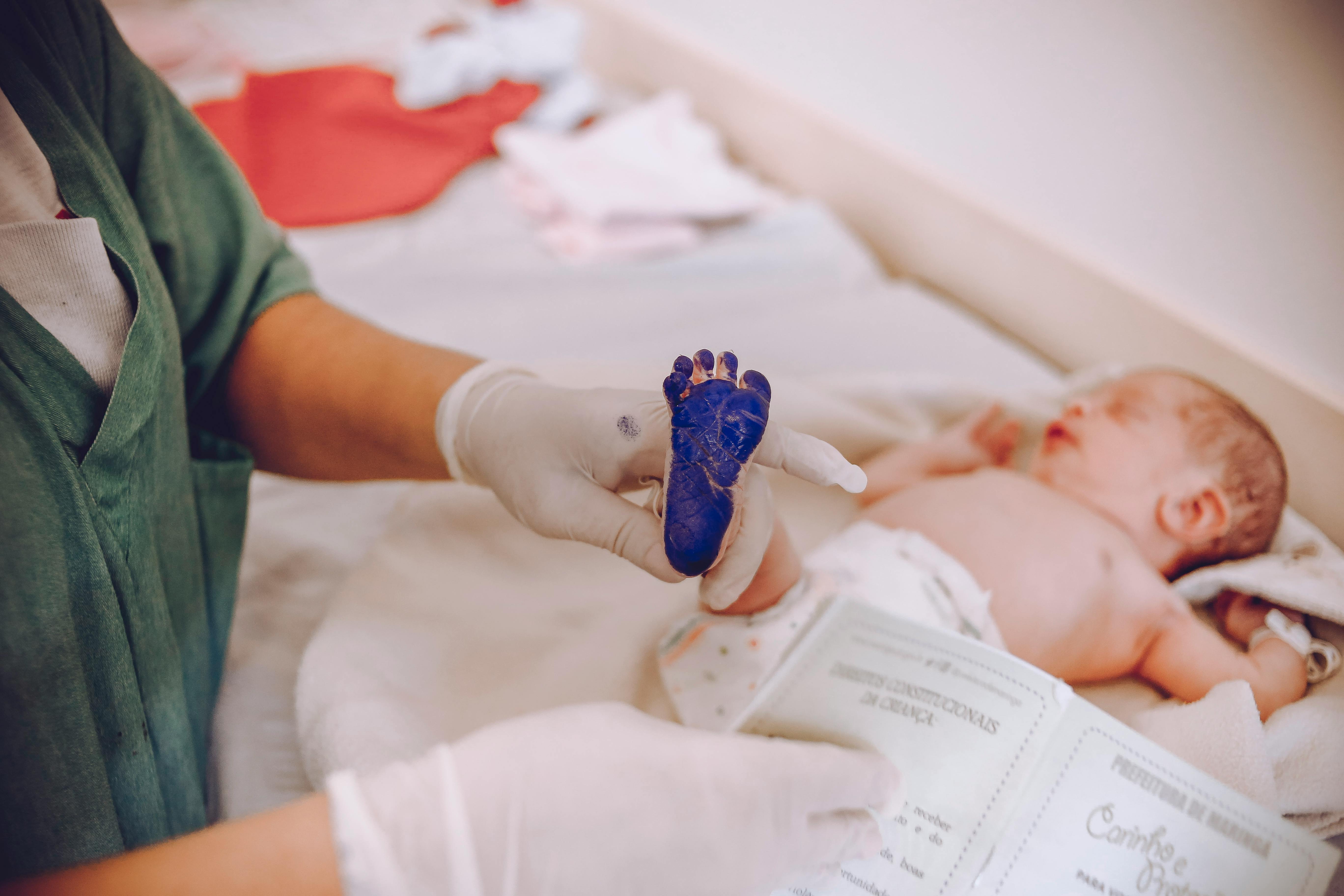 A white baby is laying on a hospital table. Their foot has blue ink on the bottom as a nurse is about to take their footprint.