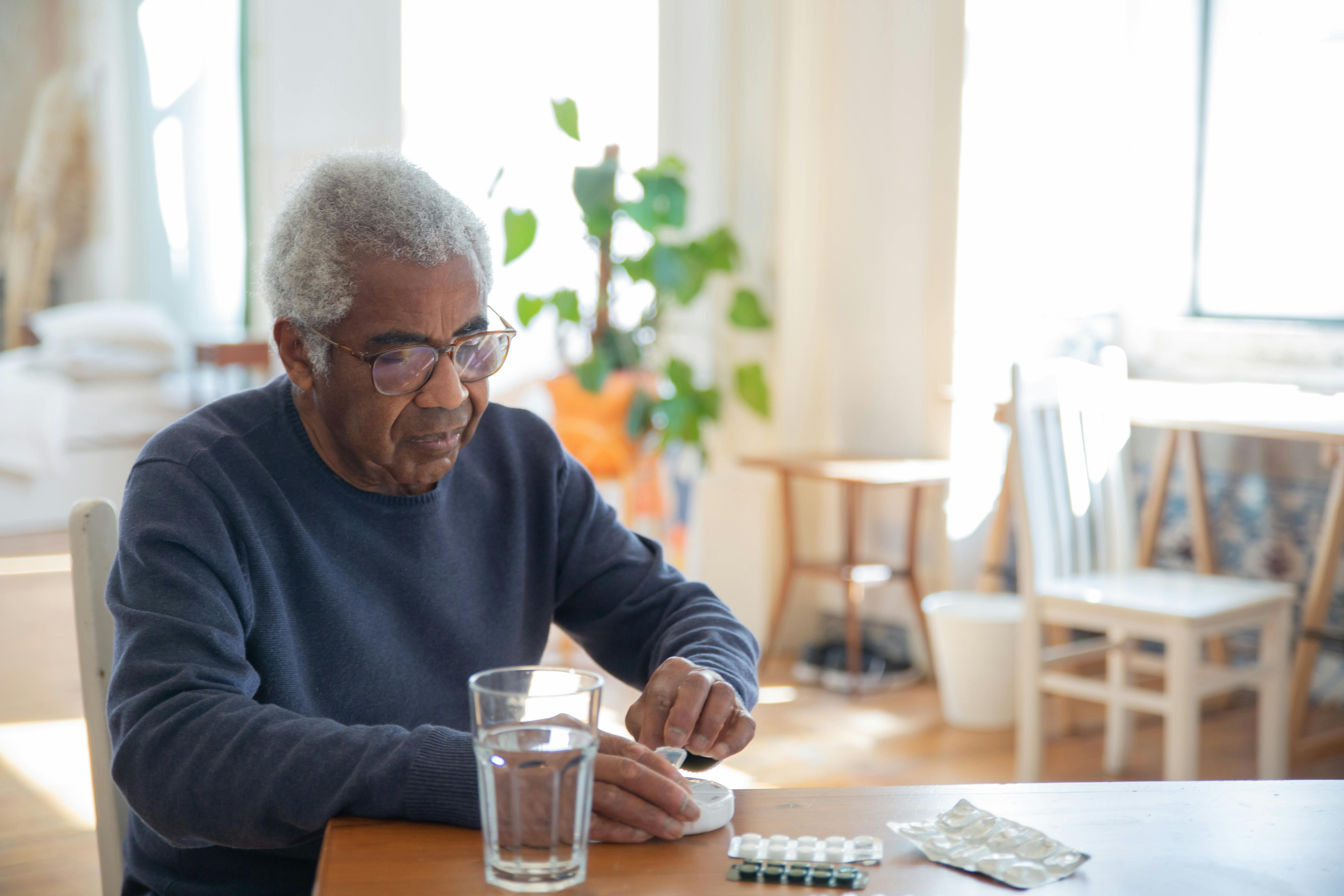 An elderly black man with gray hair sits at a kitchen table with a glass of water.