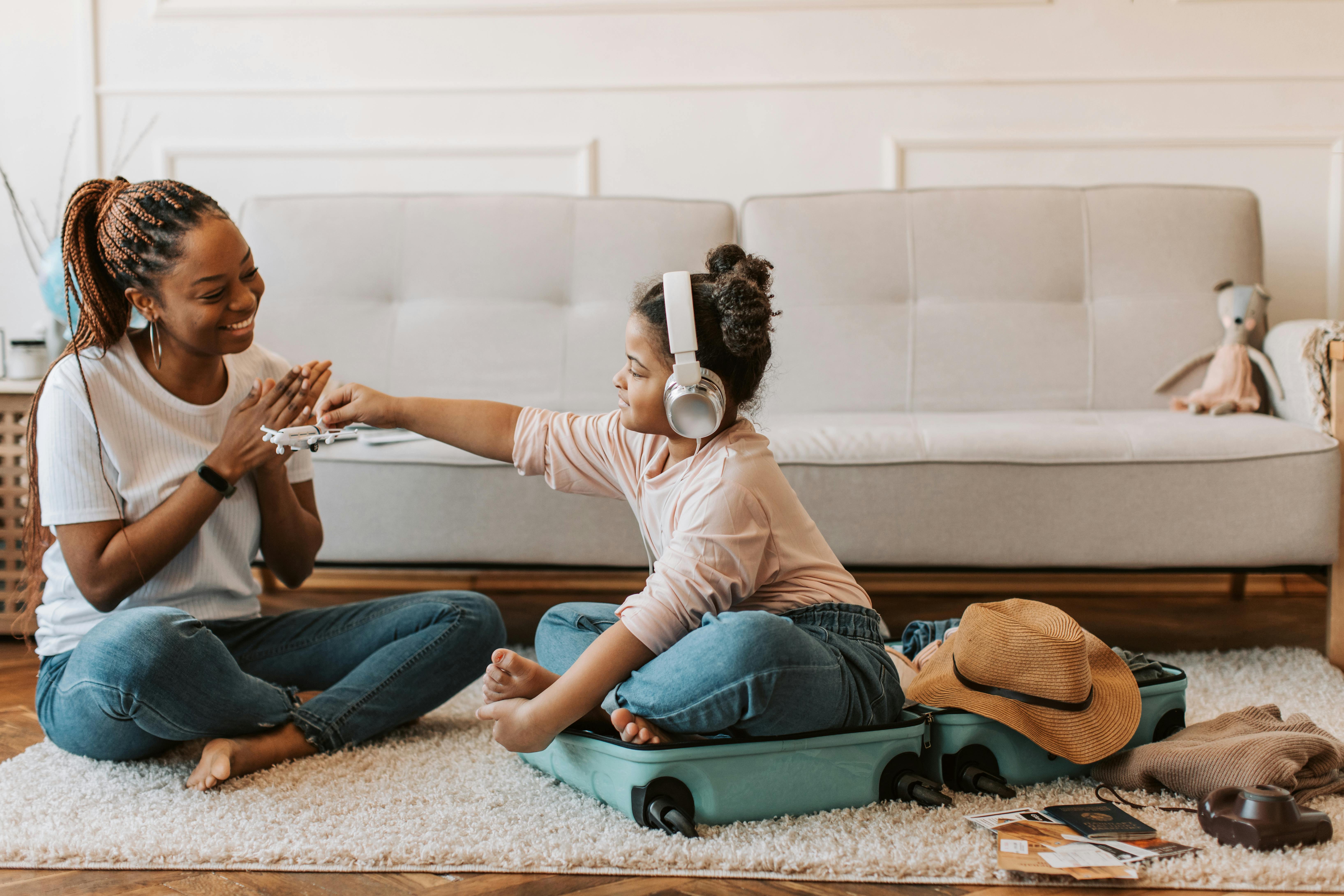 A black mother is sitting on the floor next to her daughter. She is in her suitcase playing with her mom as they pack for her visitation with her dad.
