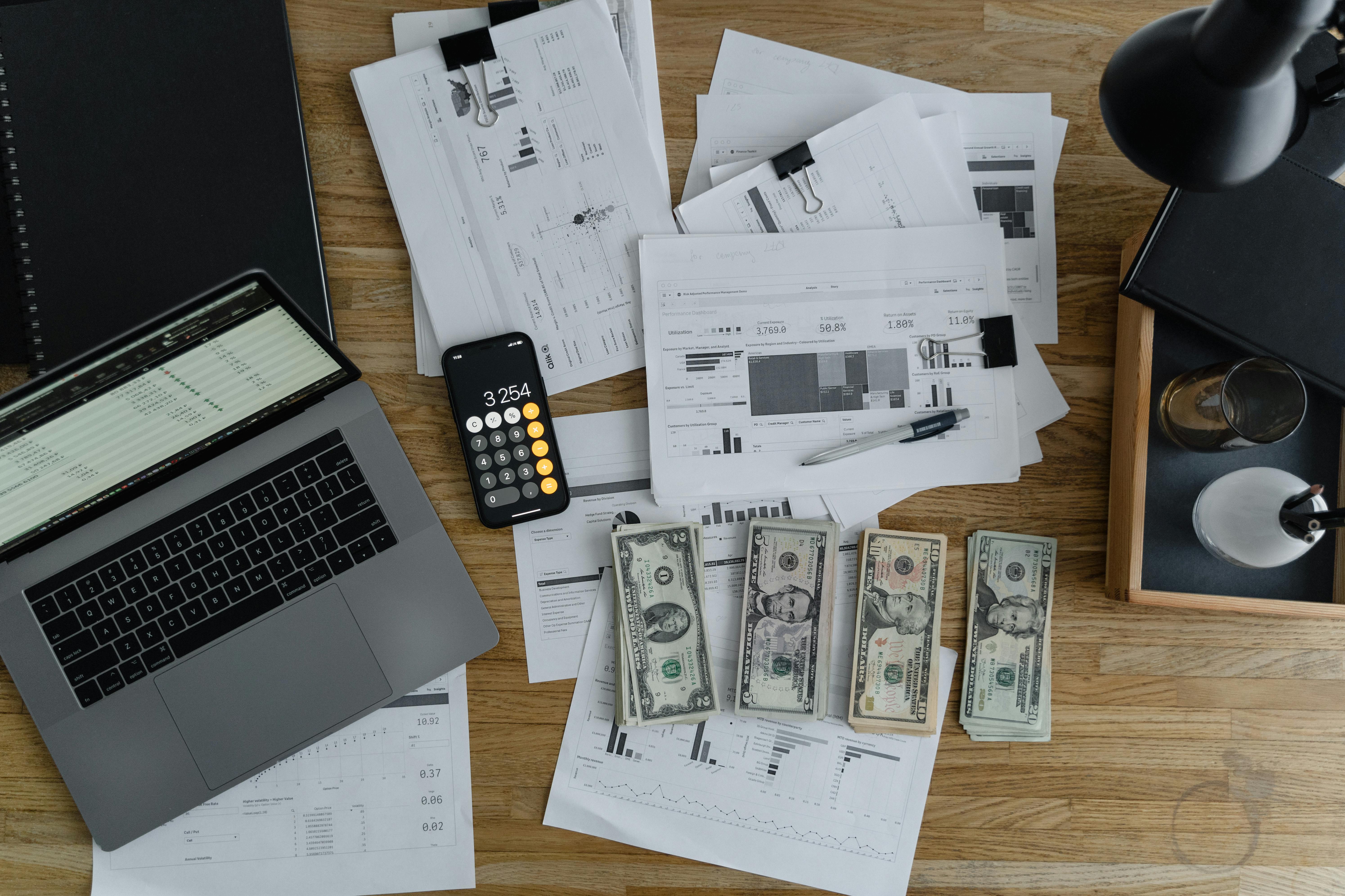 A laptop is open on a table. Next to it are multiple piles of papers, financial statements, a phone open to the calculator app, and a few piles of US cash.