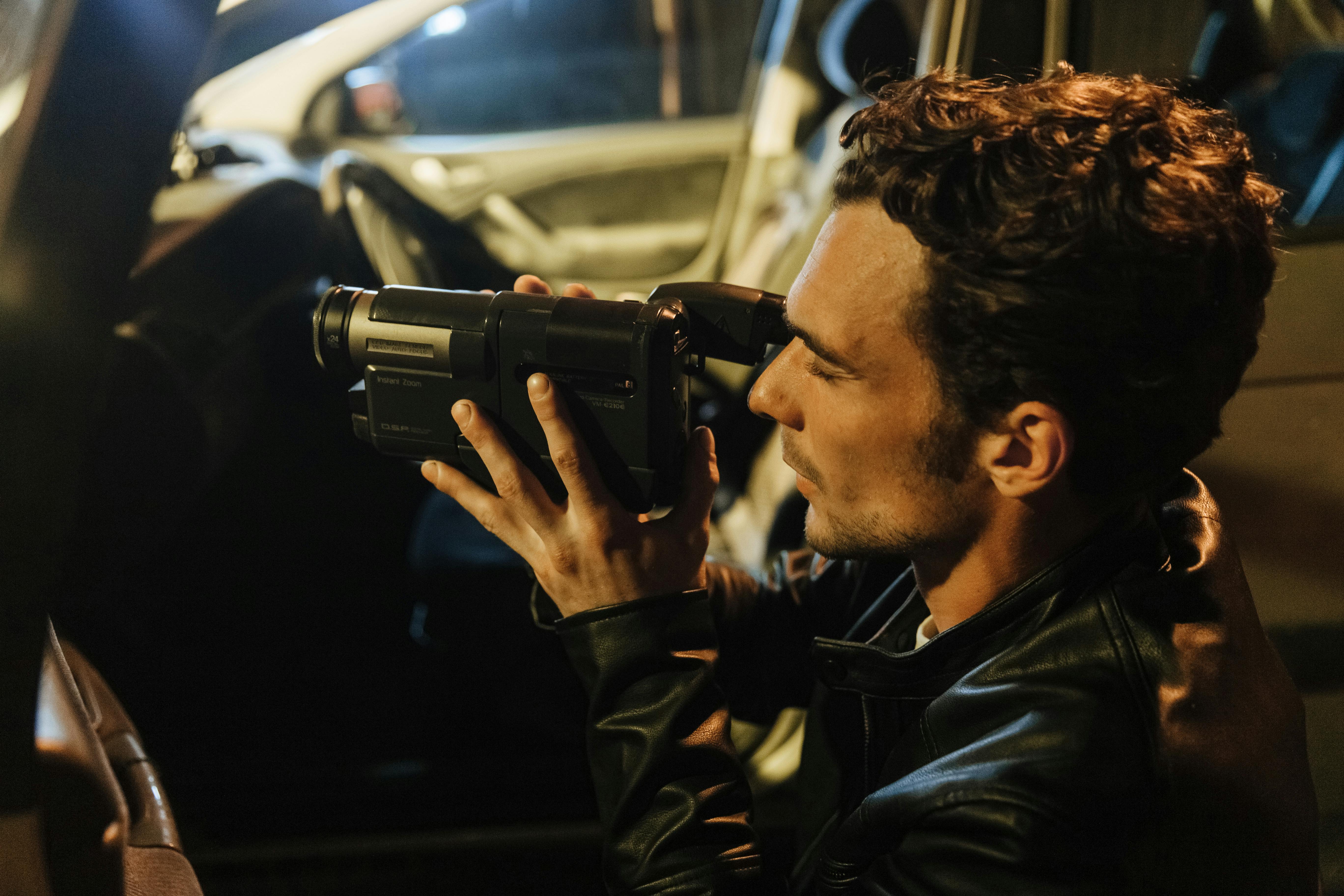 A white man sits in a car with a camcorder. He is a private investigator hired for a divorce.