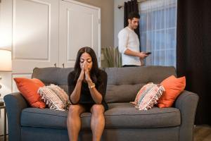 A black woman sits on a couch with her elbows on her knees. Her hands are covering her nose and mouth as her eyes are closed. Her white husband is behind her on his phone.
