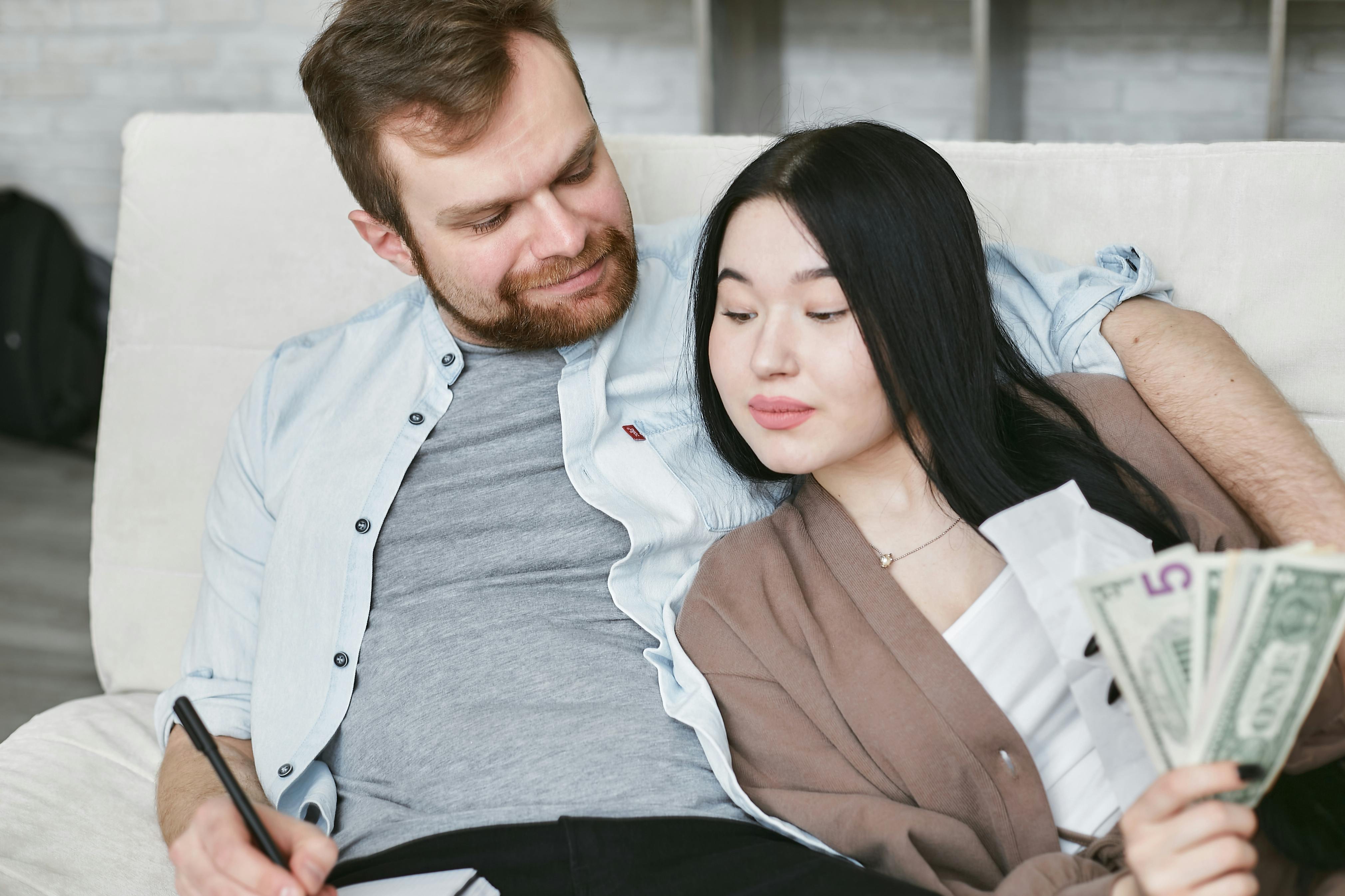A white husband and an Asian wife sit on the couch together discussing their finances. The wife is holding a fanned out handful of cash.