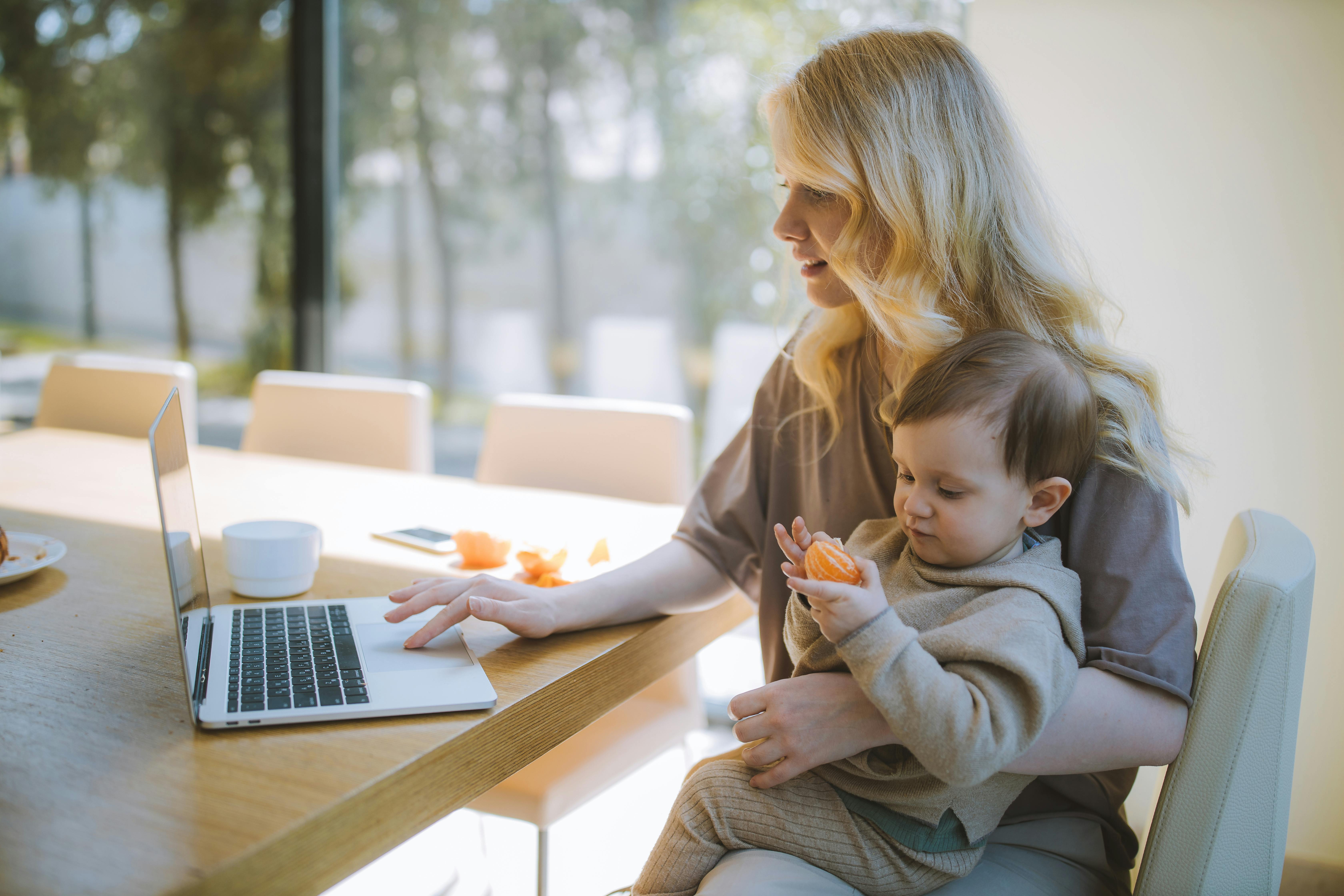 A white woman is sitting at a kitchen counter. she has her young son on her lap. He is eating a cutie as she researches divorce on her laptop.