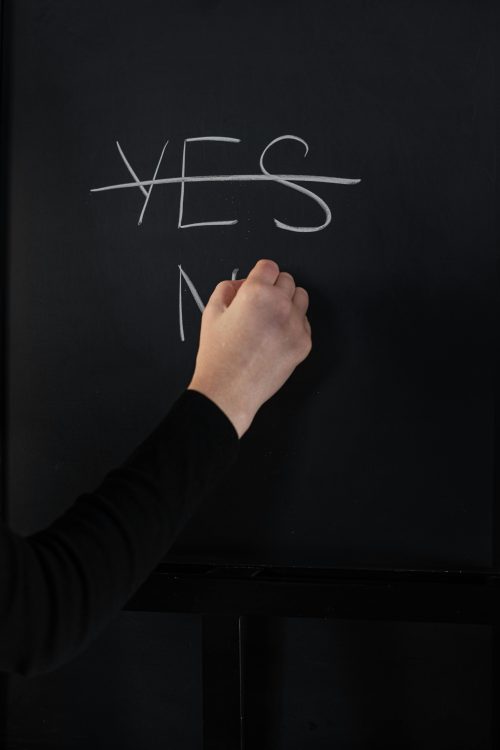 A hand has crossed out the word YES on a chalkboard and is writing the word NO beneath it.