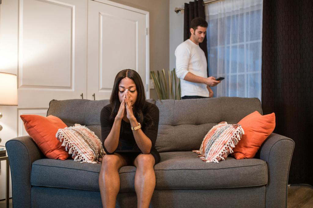 Women sitting on the couch with head in her hands frustrated about her divorce. Man on his phone in the background.